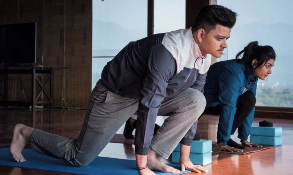 A room with a large window, a blue exercise mat, and a person performing a yoga pose.