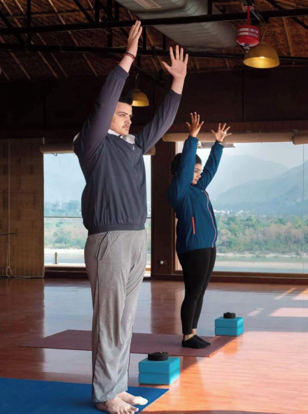 Two people practicing yoga in a room with a wooden floor, large windows, and a view of the outdoors.