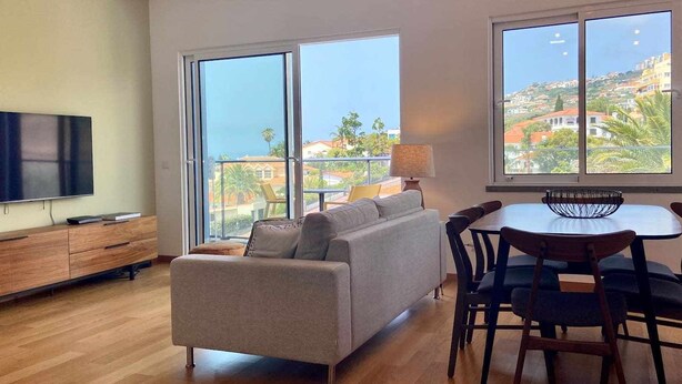 Ein modernes Wohnzimmer mit einer Couch, einem Esstisch und Stühlen, große Fenster mit Blick auf eine hügelige Landschaft.