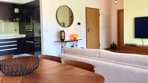 Ein modernes Wohnzimmer mit einer Couch, einem Esstisch mit Drahtkorb, einer Holzkommode, einem an der Wand montierten Fernseher und einer Küchenzeile mit dunklen Schränken und Geräten.