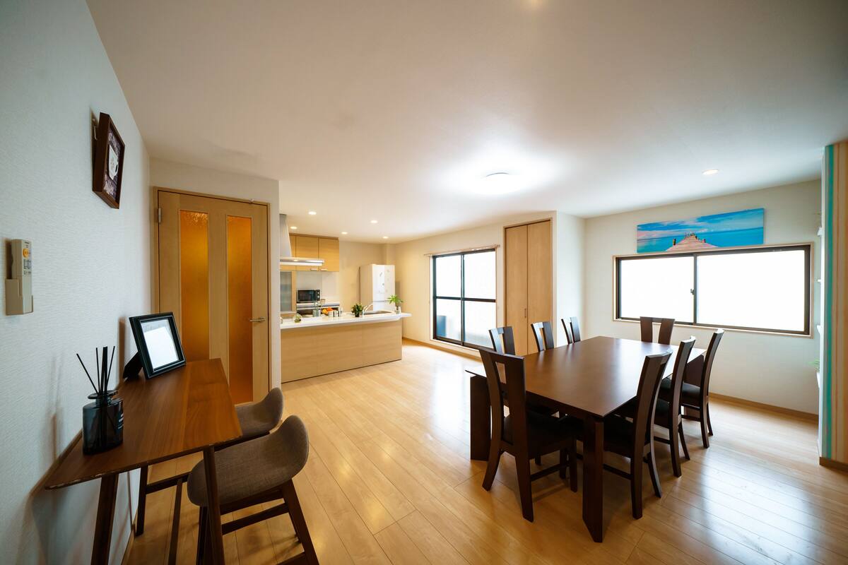 A spacious dining area with a wooden table and chairs, a kitchen in the background, and a large window with a view.