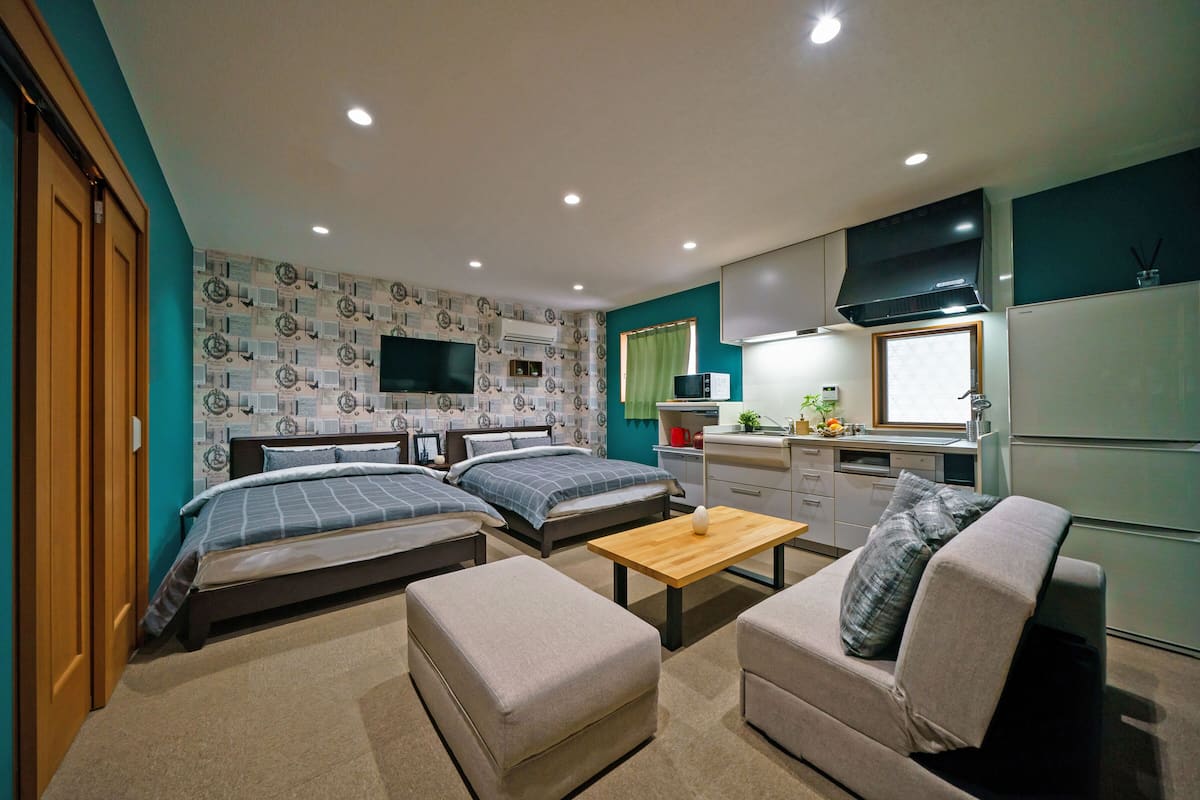 A compact, well-lit room with two beds, a sofa, a small table, and a kitchenette.