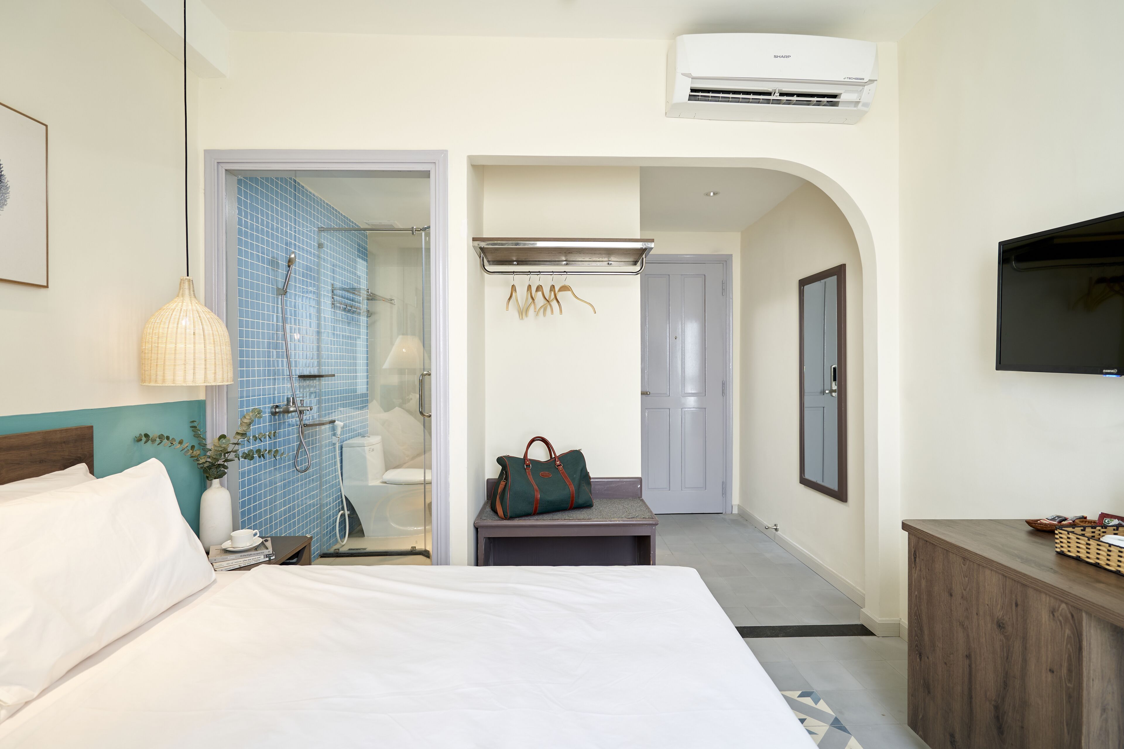 Una habitación de hotel con una cama, una mesita de noche con lámpara, un bolso colgado y un televisor sobre un mueble de madera.