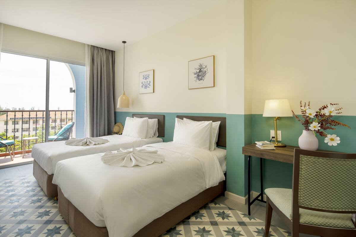 Une chambre d’hôtel avec deux lits, un balcon avec une chaise, un bureau avec une lampe et des œuvres d’art aux murs.