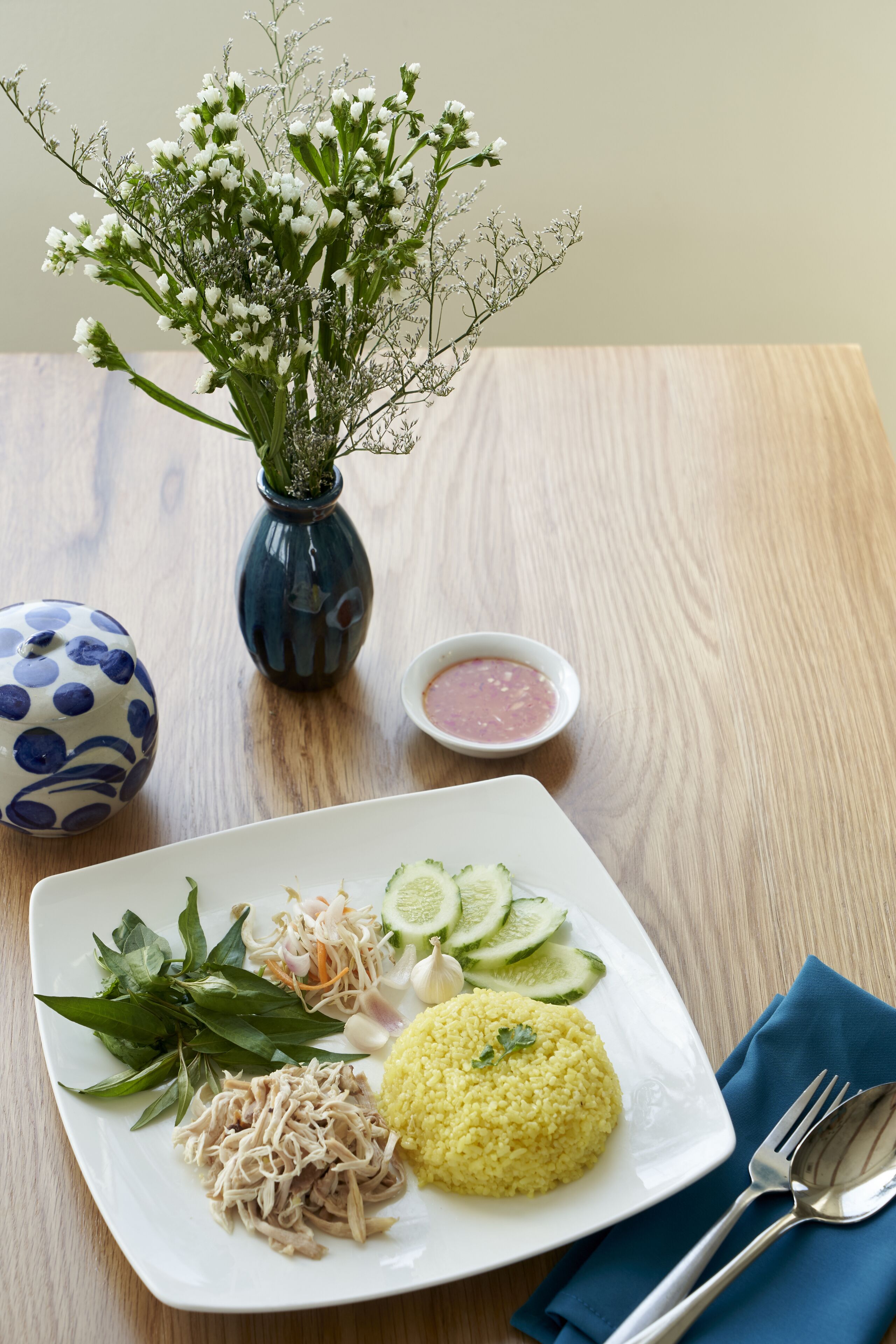 A plate with yellow rice, shredded chicken, cucumber slices, and a small bowl of sauce, accompanied by a blue vase with white flowers and a blue and white patterned vase.