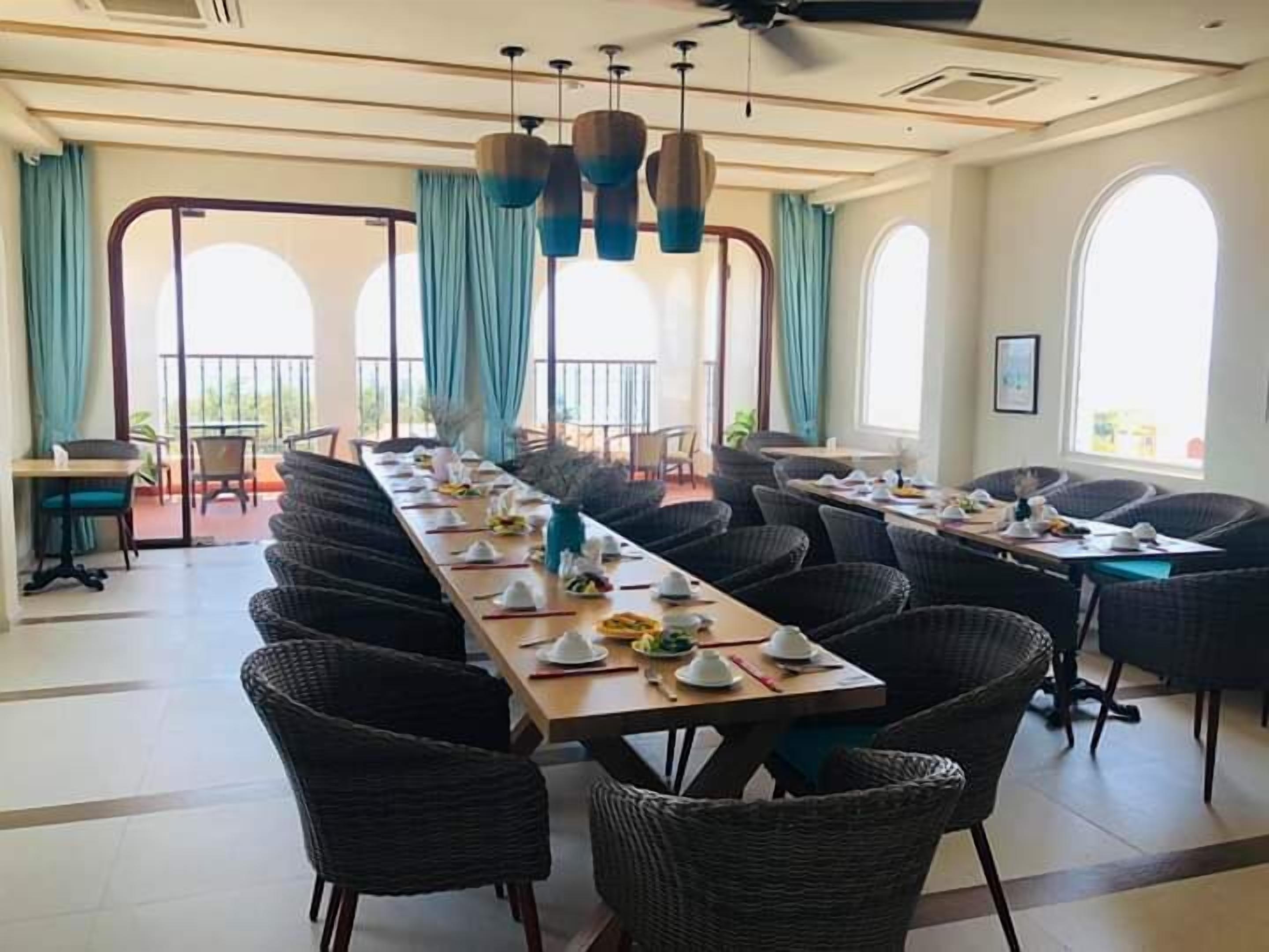 A dining area with a long table set for a meal, surrounded by wicker chairs, with large windows and curtains.