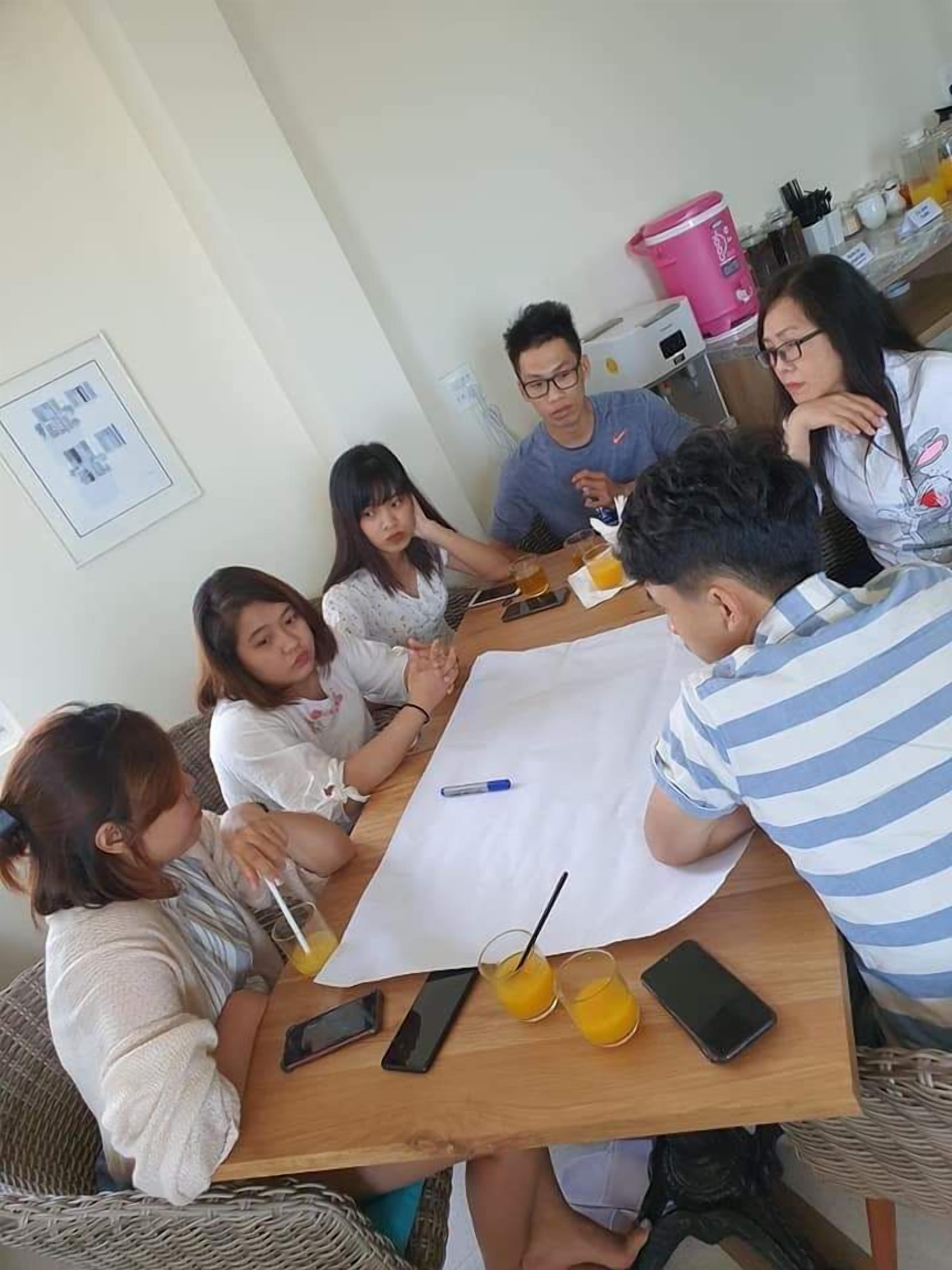 A group of people sitting around a wooden table with a white tablecloth, engaged in a discussion. There are drinks, a smartphone, and a notebook on the table.