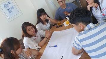 A group of people sitting around a wooden table with a white tablecloth, engaged in a discussion. There are drinks, a smartphone, and a notebook on the table.