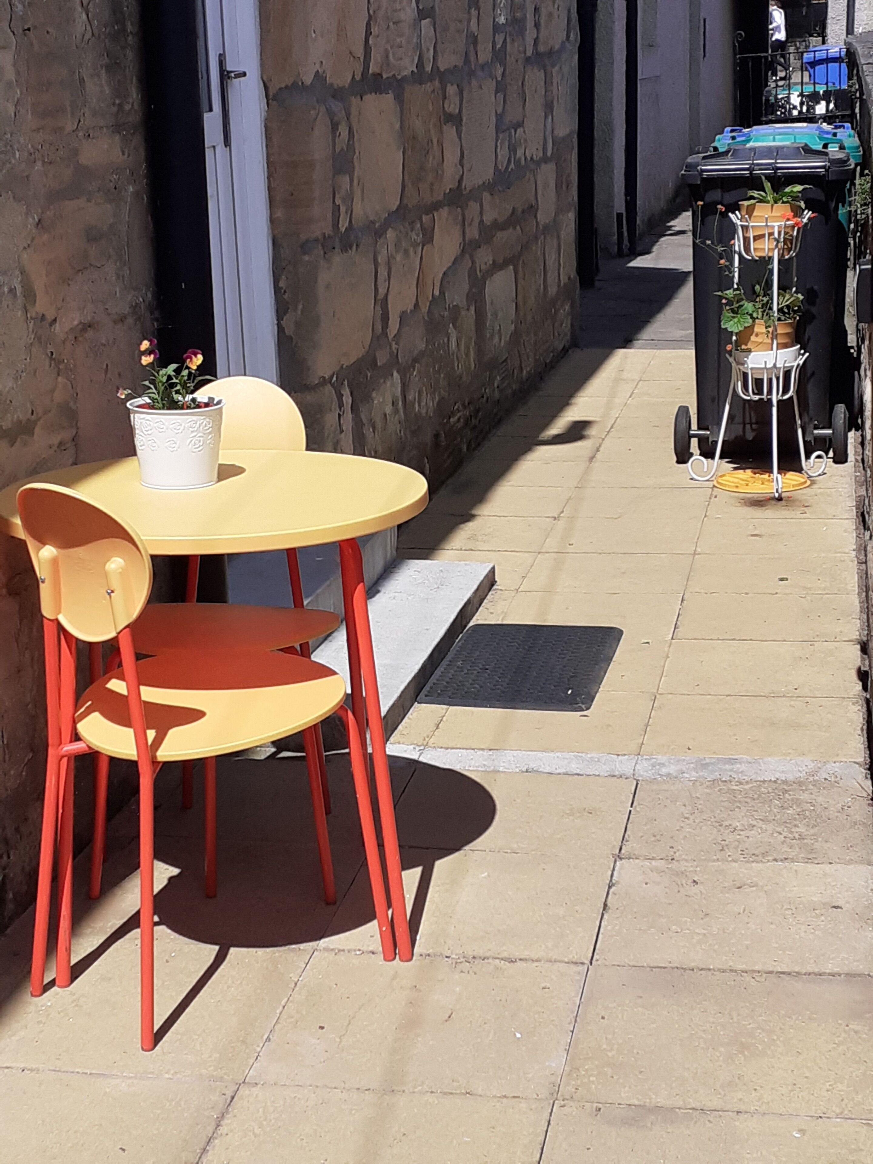 An outdoor seating area with a yellow table and red chairs, a potted plant, and a trash bin with a plant stand.