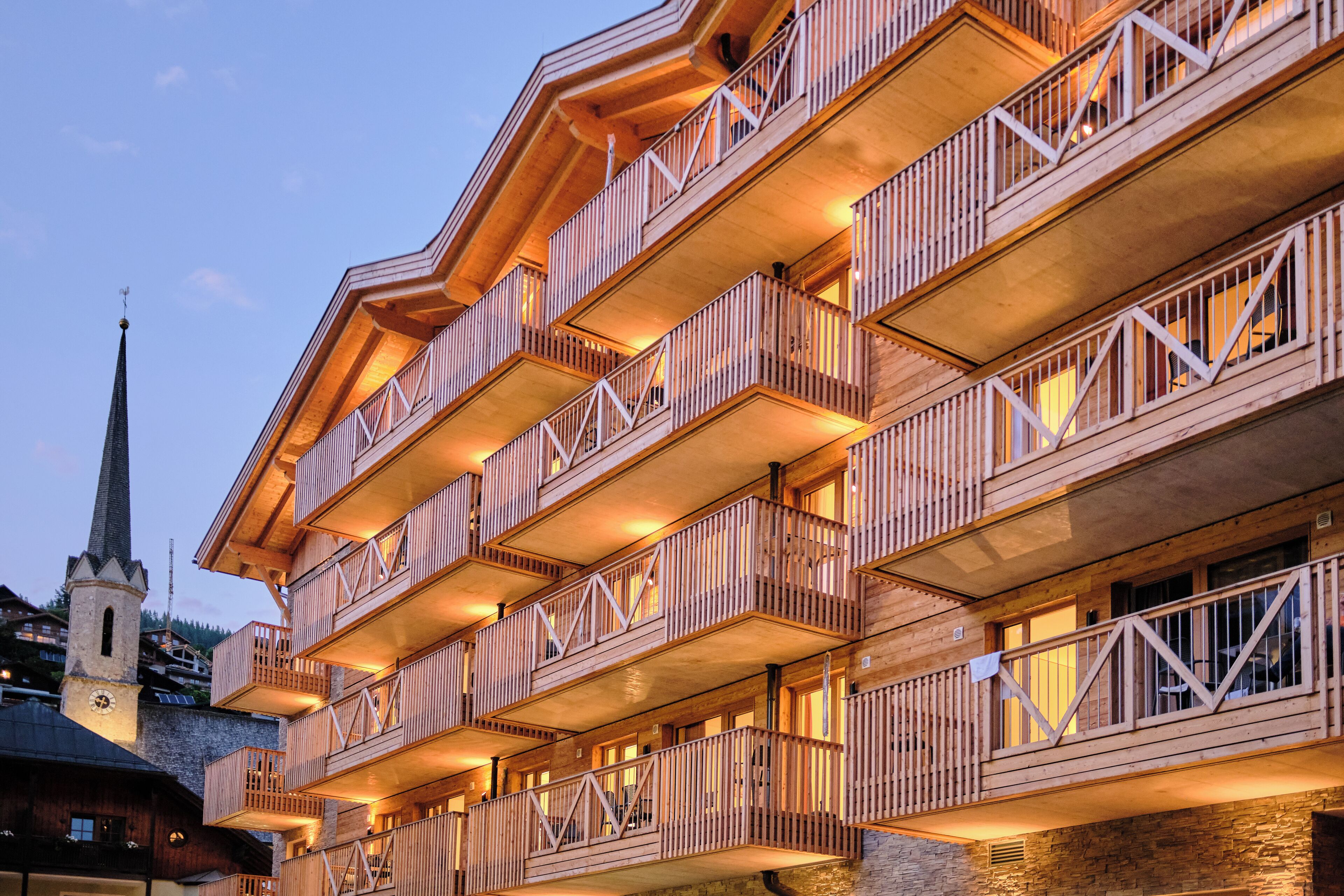 Un immeuble en bois à plusieurs étages, doté de balcons, avec le clocher d’une église en arrière-plan et un mur de pierre à sa base.