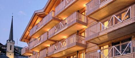Un immeuble en bois à plusieurs étages, doté de balcons, avec le clocher d’une église en arrière-plan et un mur de pierre à sa base.