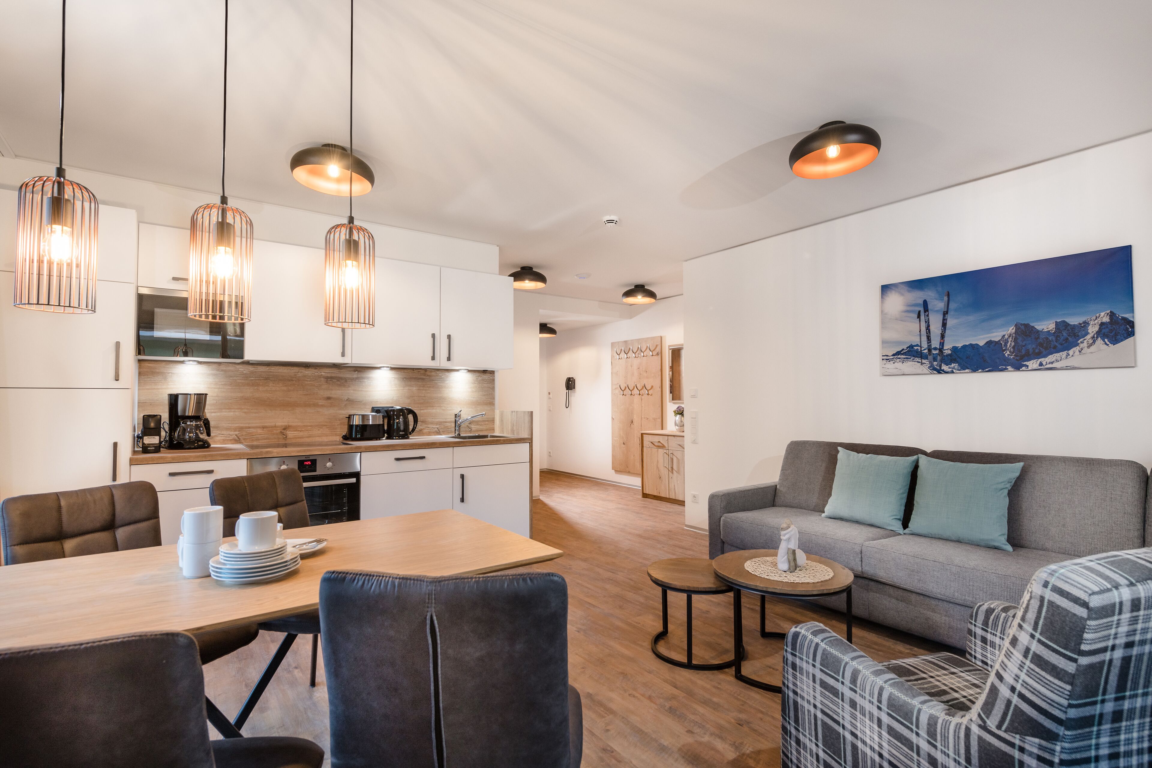 A modern kitchen with pendant lighting, a dining area with a wooden table and chairs, and a living area with a sofa and a small round table.