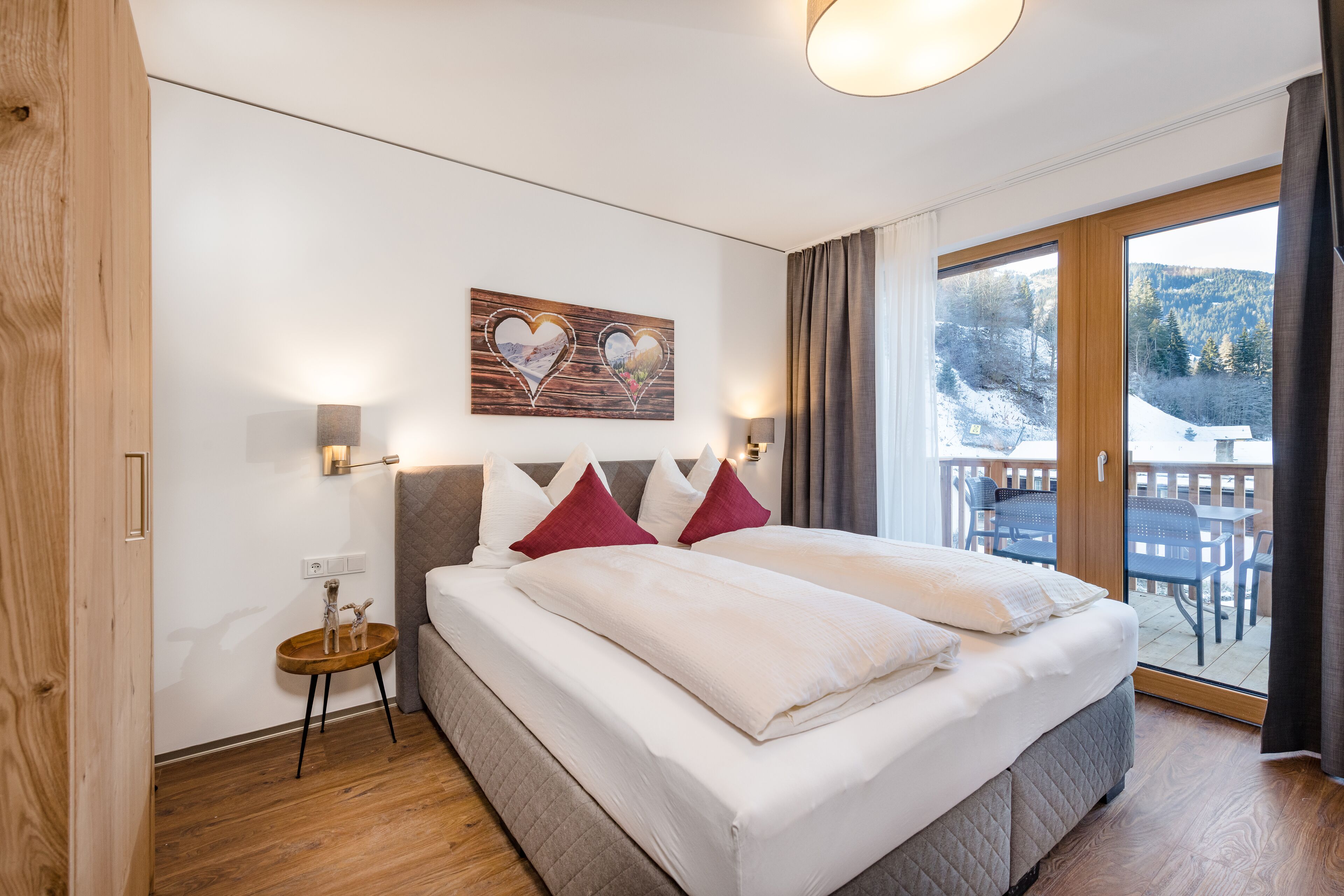Une chambre d’hôtel avec un grand lit, un plancher en bois, un balcon offrant une vue sur les montagnes et un tableau représentant des cœurs accroché au mur.