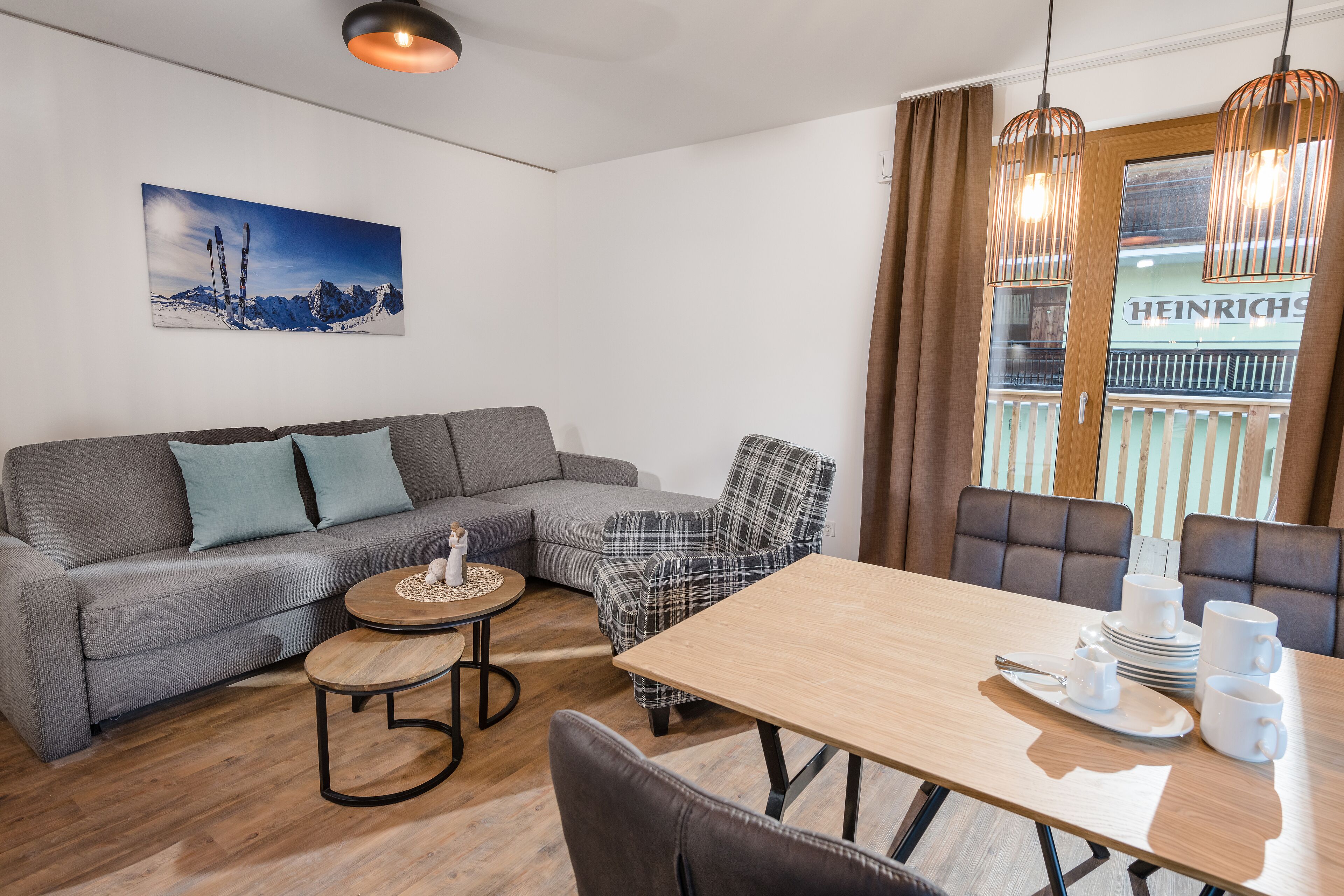 En moderne stue med en grå sofa, en ternet lænestol, et træspisebord dækket med hvide kopper og tallerkener, samt et maleri af et bjerglandskab på væggen.