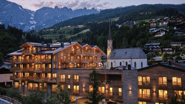 A multi-story wooden building with balconies, a church, and a mountain backdrop. - AlpenParks Hotel & Apartment Hochkönig (Muehlbach Am Hochkoenig)