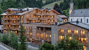 A multi-story wooden building with balconies and large windows, surrounded by greenery and a church. - AlpenParks Hotel & Apartment Hochkönig (Muehlbach Am Hochkoenig)