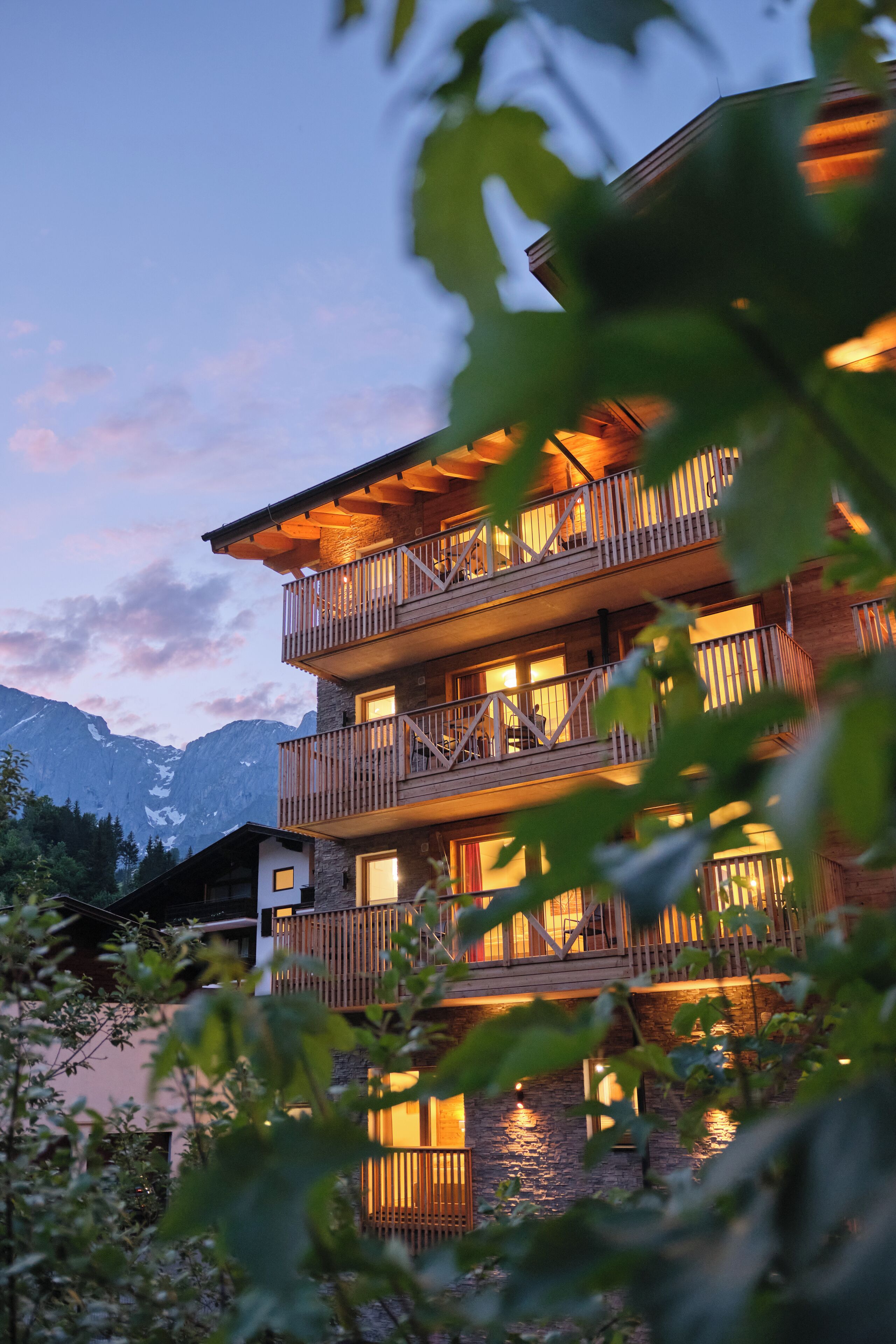 A multi-story building with balconies, surrounded by greenery and mountains in the background.