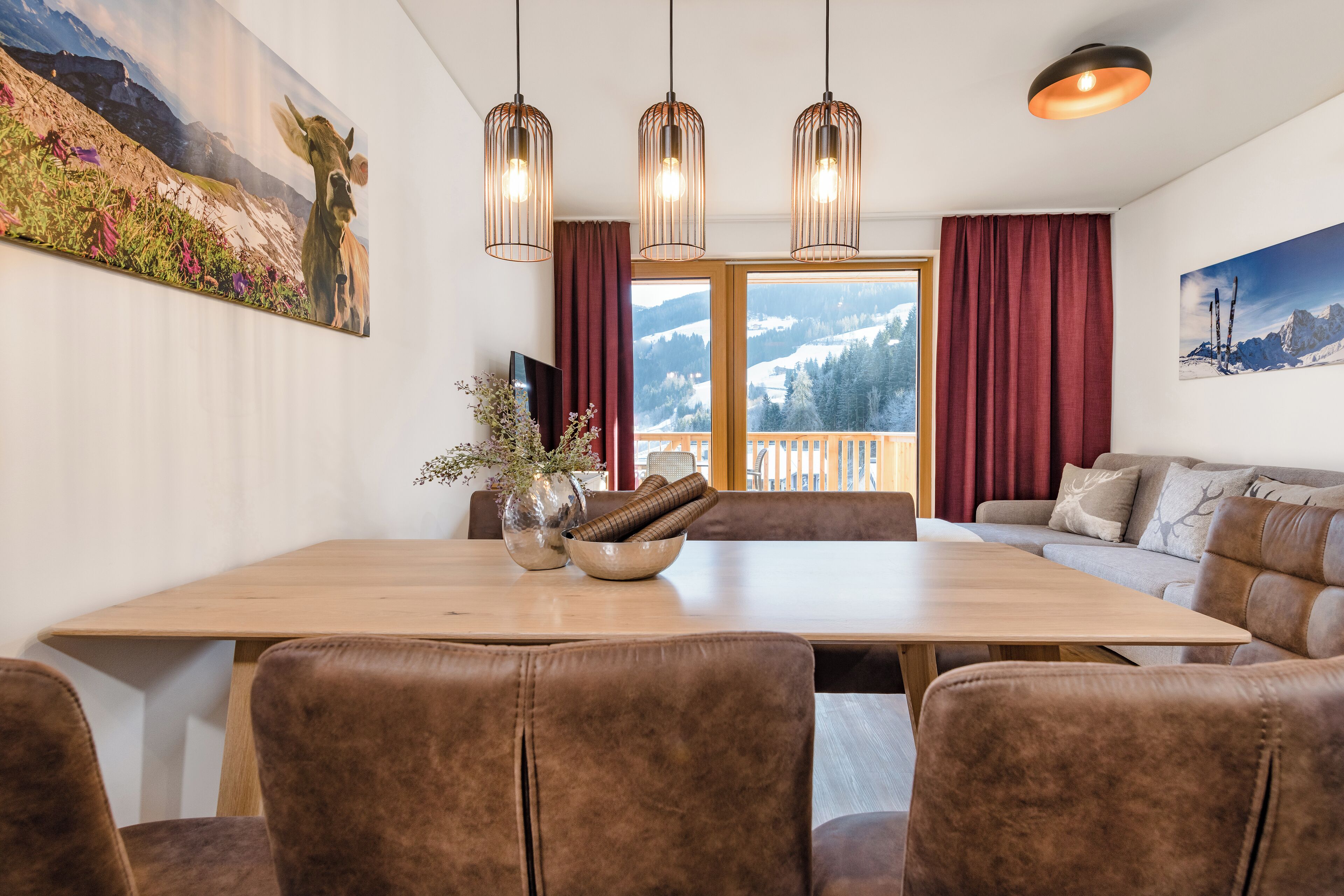 A dining area with a wooden table, brown upholstered chairs, and a sofa with cushions. There are three pendant lights above the table, and a window with curtains on the right side.
