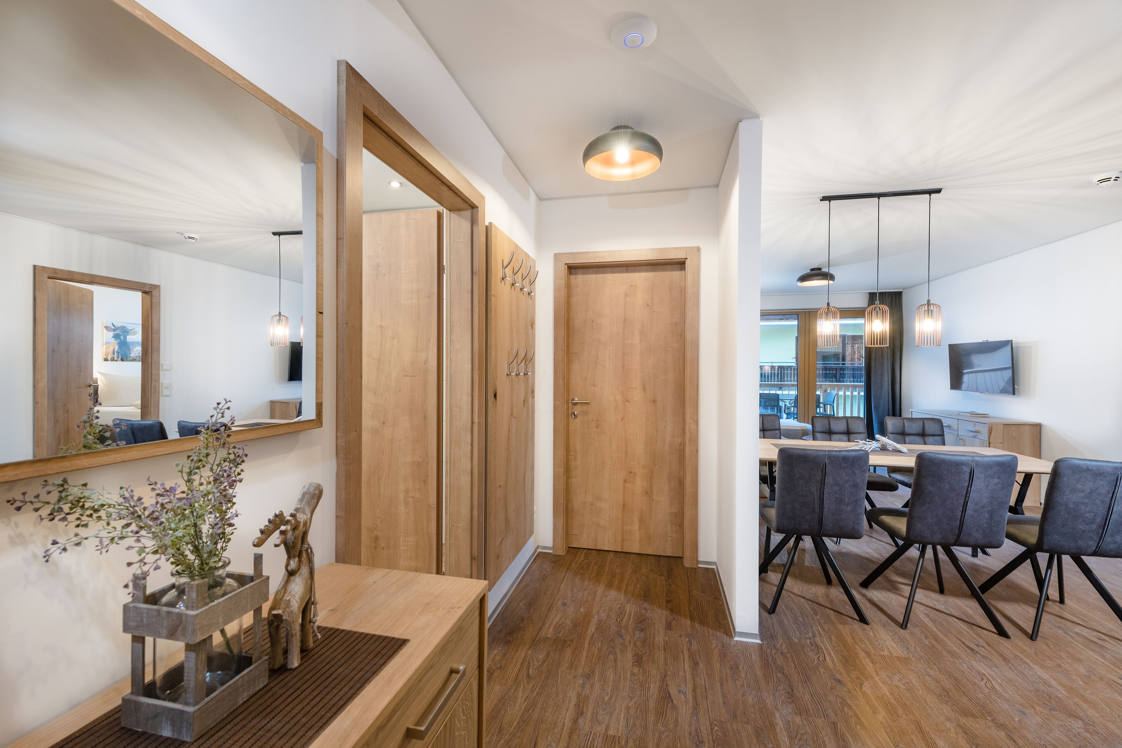 Moderni hotellihuone, jossa on puulattia, ruokailutila pöytineen ja tuoleineen, suuri peili ja ovi, joka johtaa toiseen huoneeseen.