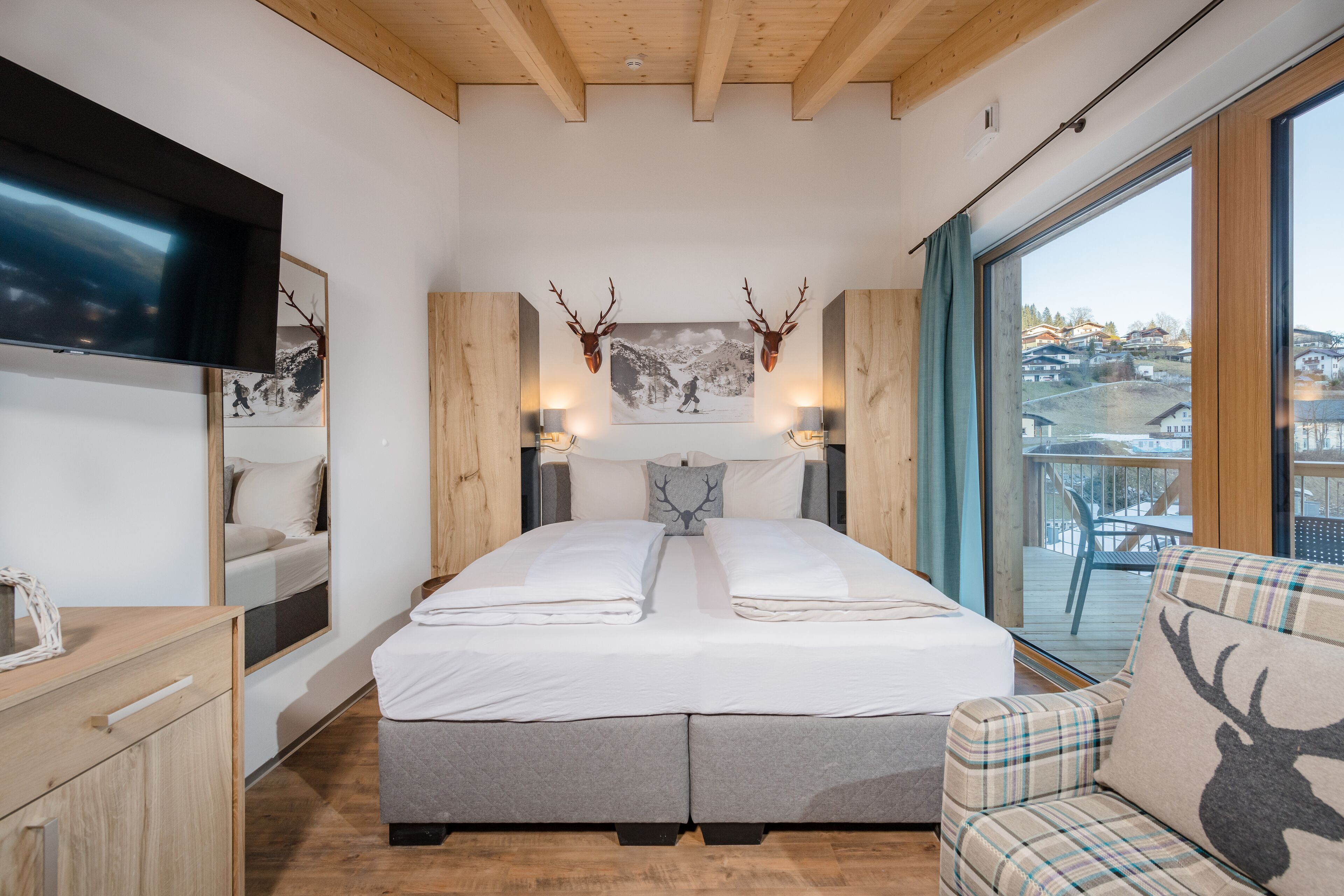 A bedroom with a large bed, wooden headboard, mounted deer antlers, a television, and a view of a balcony with outdoor seating.