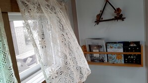 Ein Zimmer mit weißem Spitzenvorhang, einem Holzbodenregal mit Büchern und einem dekorativen Gegenstand sowie einem Fenster mit Jalousien.