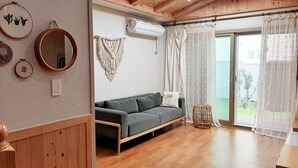 Ein gemütliches Zimmer mit Holzbalkendecke, einem grauen Sofa, einem Wandbehang aus Makramee und einem großen Fenster mit durchsichtigen Vorhängen.