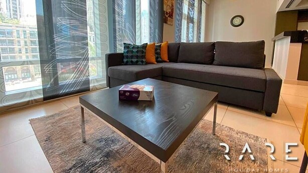 A modern living room with a dark sofa, a coffee table, and large windows with curtains. - Stylish 1 Bed apartment in Downtown Dubai (Dubai)