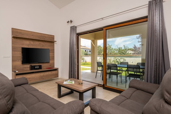 A living room with a flat-screen TV, wooden TV stand, and a sliding glass door leading to an outdoor patio with a table and chairs. - Holiday house Evida in Poreč (Poreč)