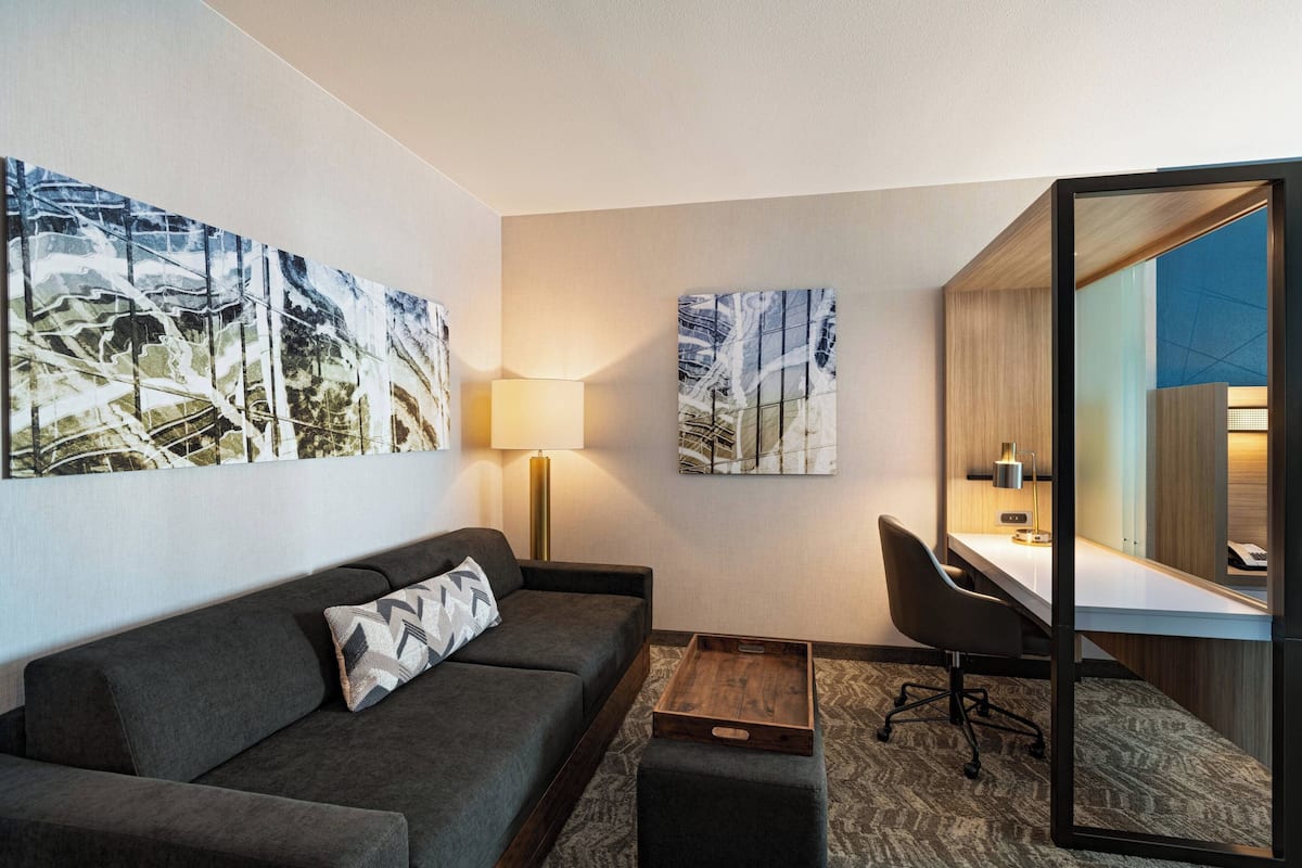 A modern hotel room with a grey sofa, a desk with a chair, and two large abstract paintings on the wall.
