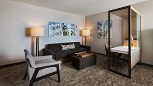 Ein Hotelzimmer mit einer Couch, einem Sessel, einem Schreibtisch und zwei Lampen. Zwei Bilder hängen an der Wand und es gibt einen Spiegel.