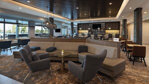 Eine moderne Hotellobby mit einer Eckcouch, Sessel, einem kleinen runden Tisch und einer Bar mit Sitzgelegenheiten.