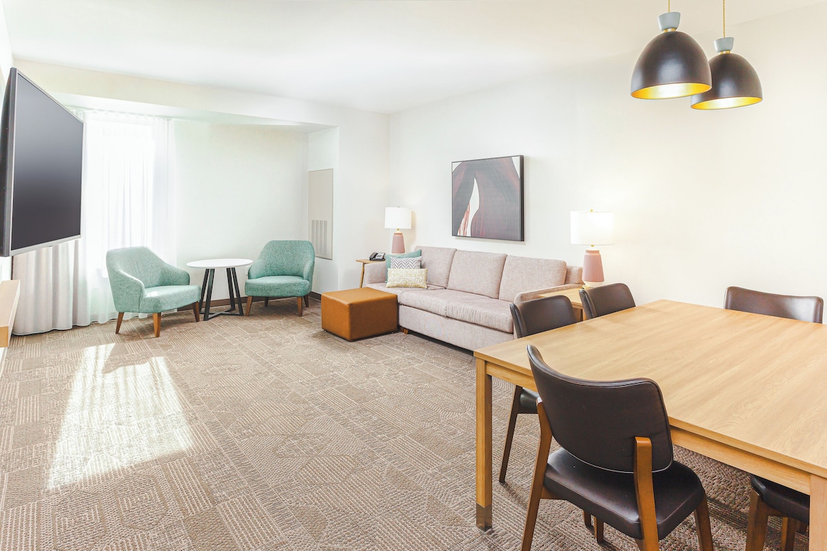 Quarto de hotel com sofá, duas poltronas, uma mesa de jantar de madeira e televisão.