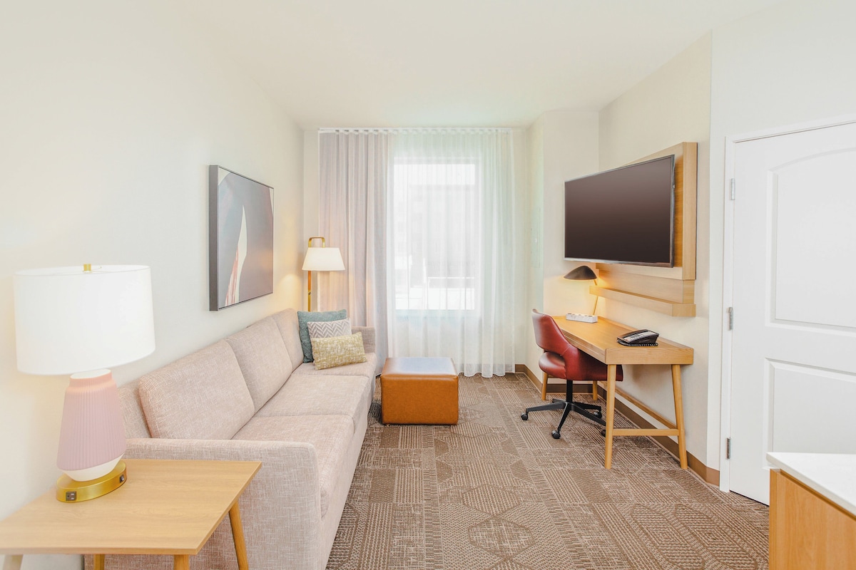 Quarto de hotel com sofá, mesa pequena, escrivaninha, cadeira, TV e janela com cortinas.