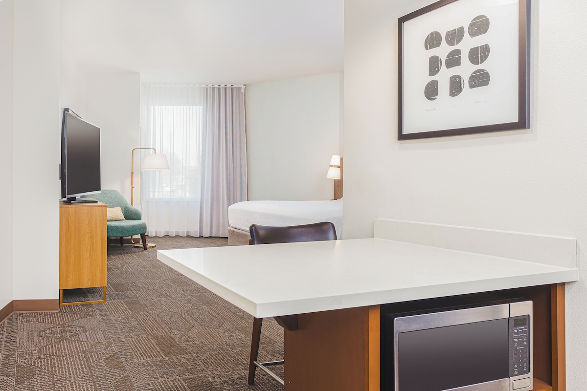 Quarto de hotel com televisão de tela plana, escrivaninha com micro-ondas, cadeira, luminária e uma janela grande com cortinas.