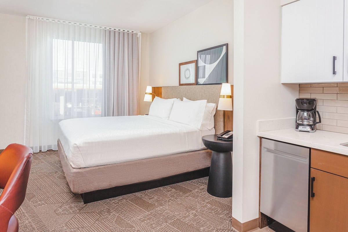 Quarto de hotel com uma cama, uma mesinha redonda com abajur, cafeteira e vista para a cidade pela janela.