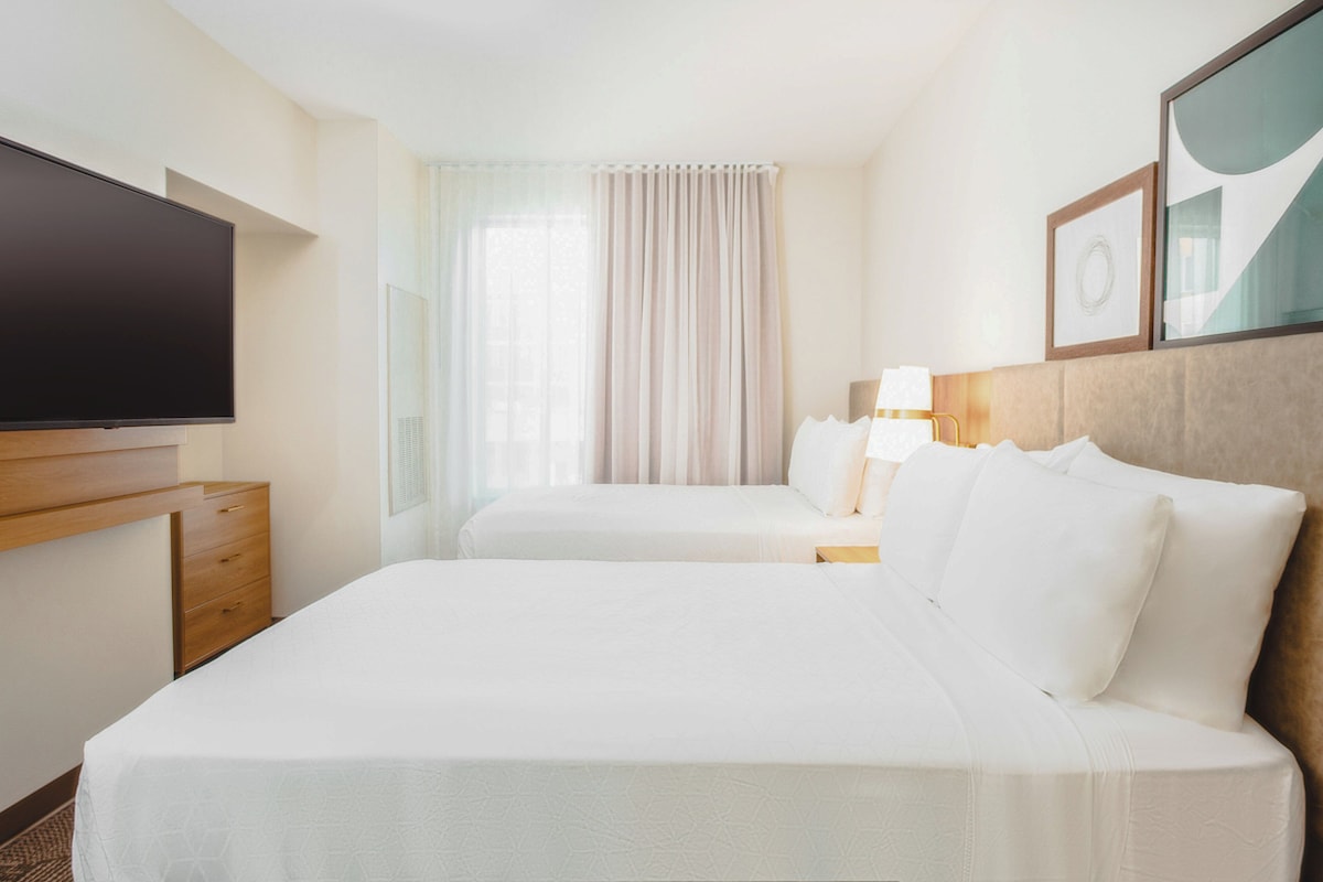 Quarto de hotel com duas camas, televisão de tela plana, mesa de cabeceira de madeira, luminária e uma janela grande com cortinas.