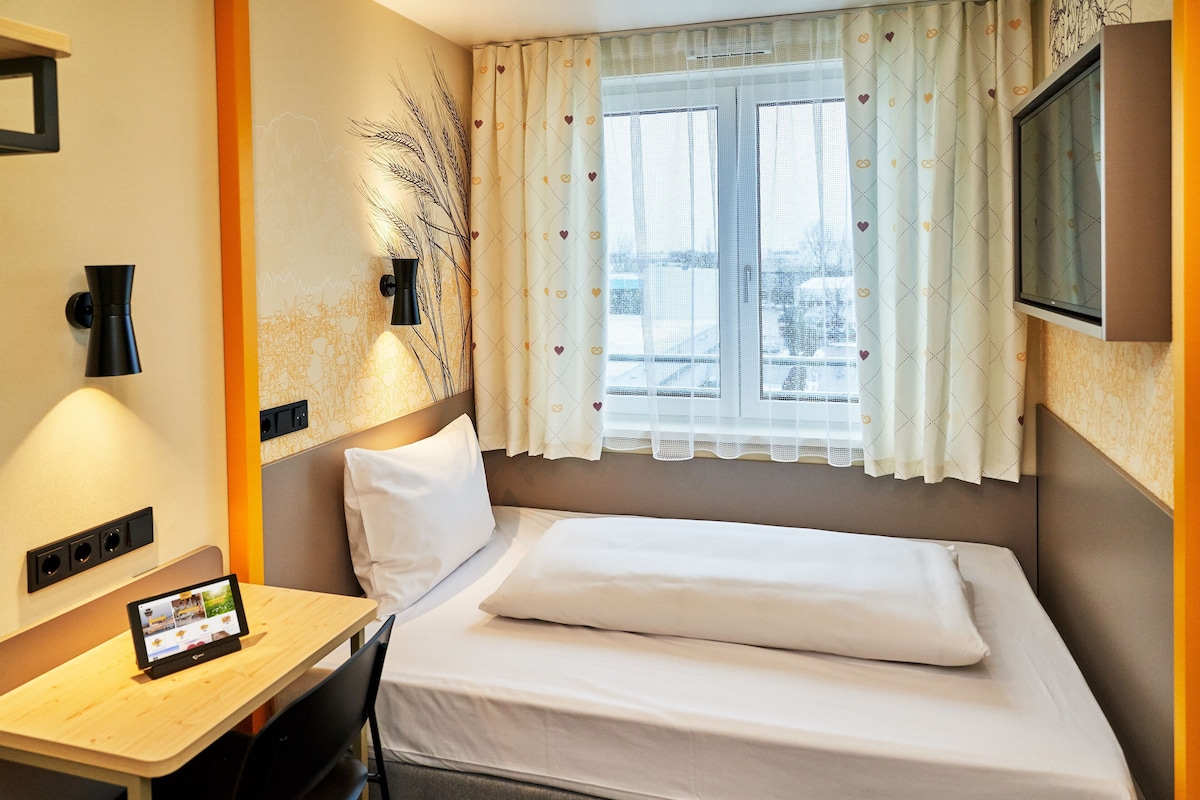 Une chambre d’hôtel avec un lit, un bureau équipé d’une tablette, une télévision et une fenêtre avec des rideaux.