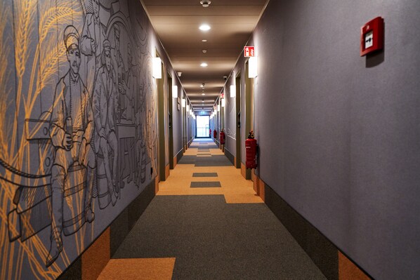 Un long couloir étroit aux murs décorés de motifs, avec un extincteur et une alarme incendie sur le côté droit.