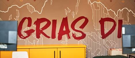 A room with a yellow desk, a white chair, and a wall with a large red text "GRIAS DI" and a line drawing of a mountain range.