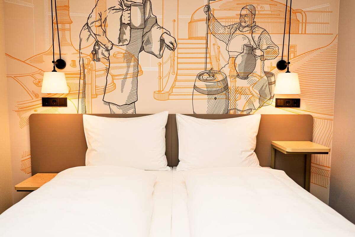 Une chambre d’hôtel avec un grand lit, deux lampes de chevet et une fresque murale représentant deux personnages.