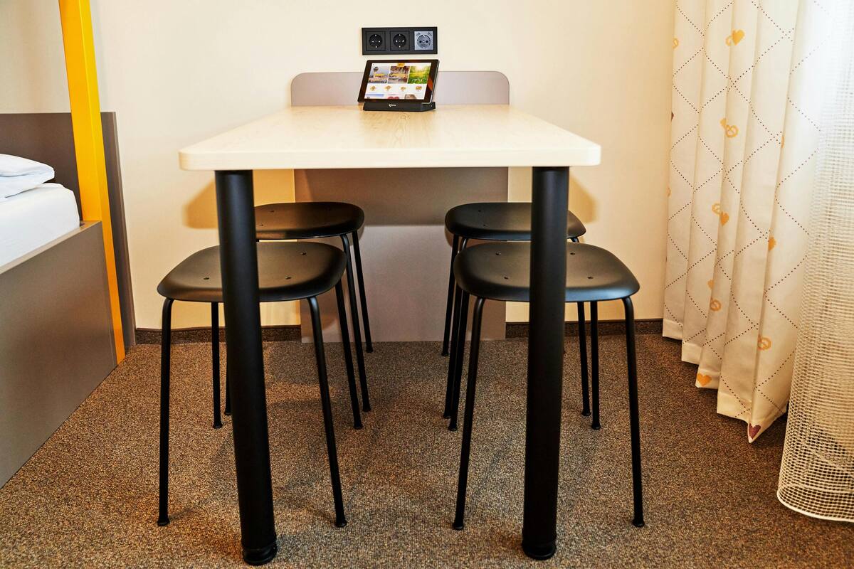 Une petite chambre d’hôtel avec une table blanche, deux chaises noires, une table de chevet avec une lampe et un rideau orné d’un motif en forme de cœur.