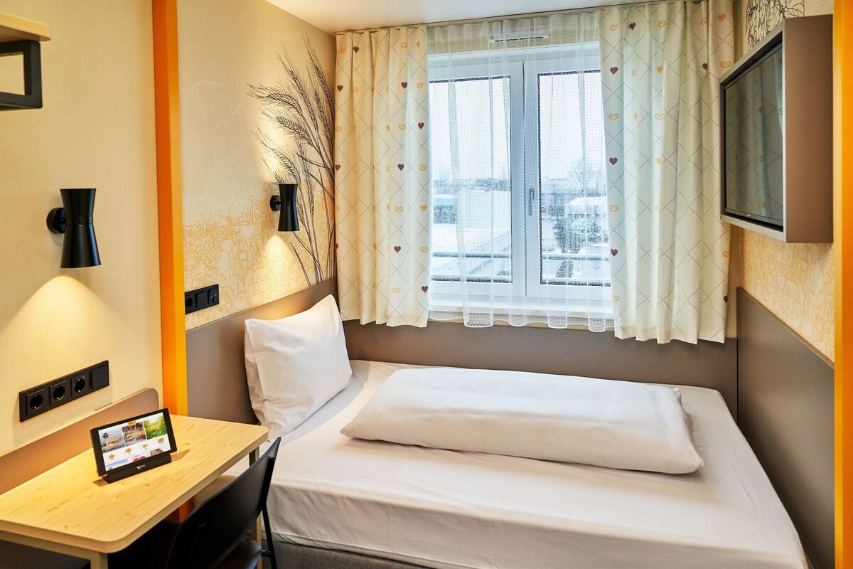 Une chambre d’hôtel avec un lit, un bureau équipé d’une tablette, une télévision et une fenêtre avec des rideaux.