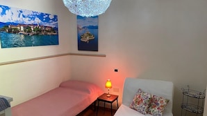Ein kleiner, gemütlicher Raum mit einem Einzelbett, einem Sessel, einem Tisch mit Lampe und einem Wandregal.