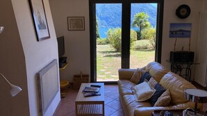 Ein Wohnzimmer mit einer Couch, einem Couchtisch und einem Fernseher. Ein Fenster bietet Blick auf einen Garten und Berge.