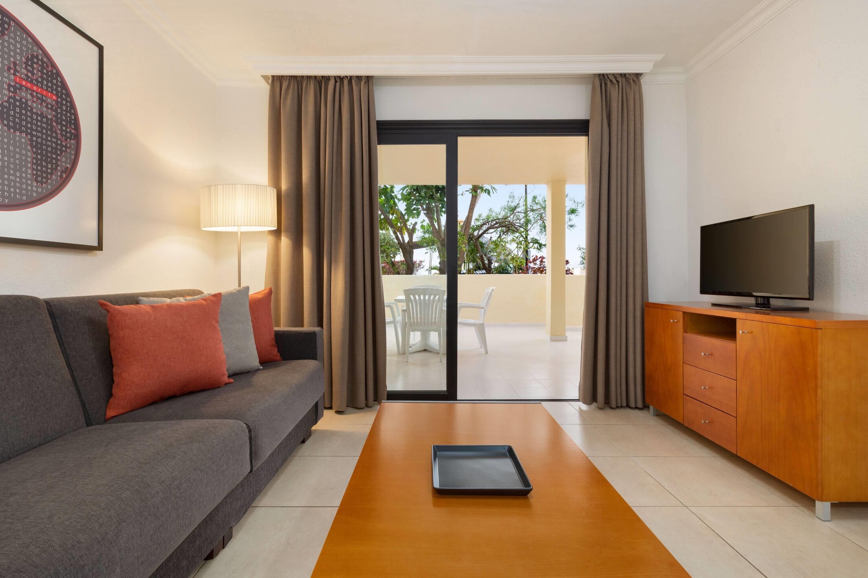 A modern living room with a grey sofa, a wooden coffee table, a television on a wooden cabinet, and a sliding glass door leading to an outdoor area with a white chair and table.
