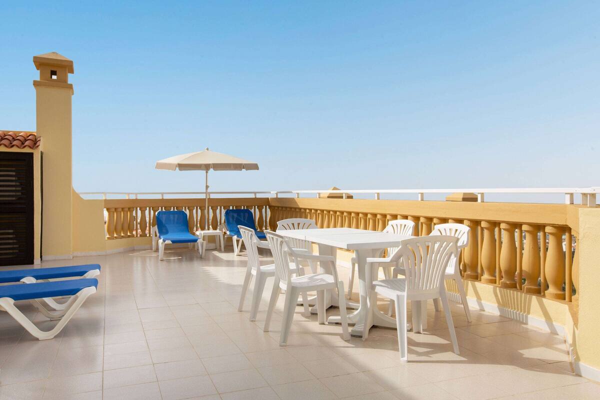 Eine Terrasse mit weißen Plastikstühlen und -tischen, einem blauen Liegestuhl und einem beigen Sonnenschirm.