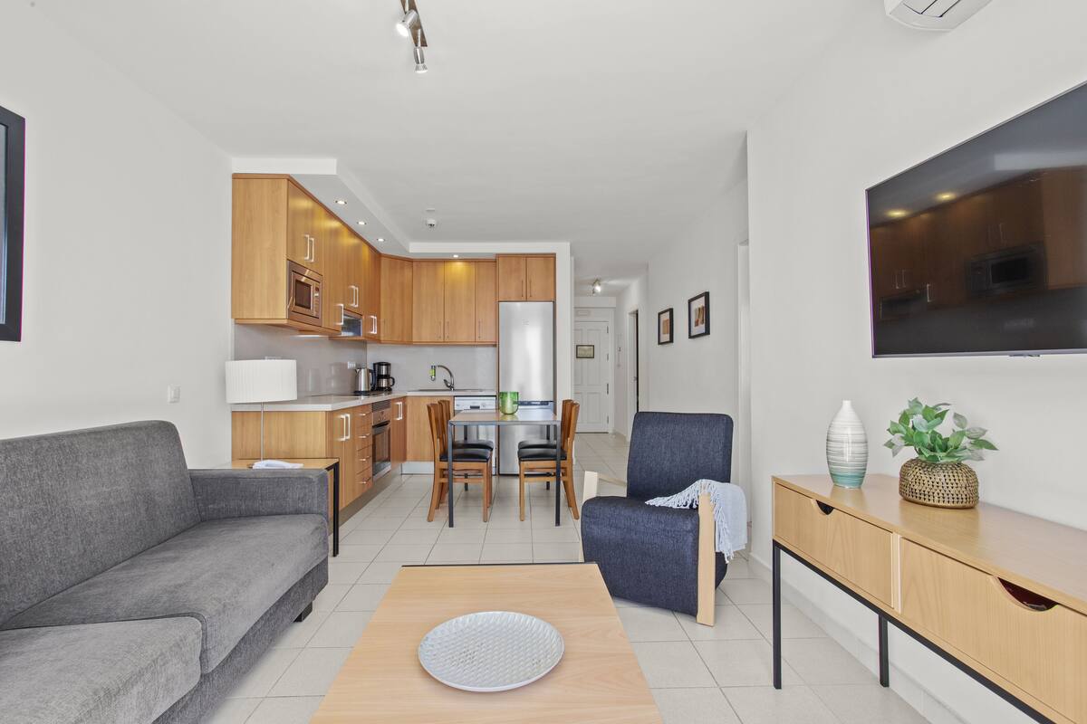 Ein modernes Wohnzimmer mit einem grauen Sofa, einem Holztisch und einem blauen Sessel. Im Hintergrund ist eine Küche mit Holzschränken und einem Edelstahlkühlschrank zu sehen.