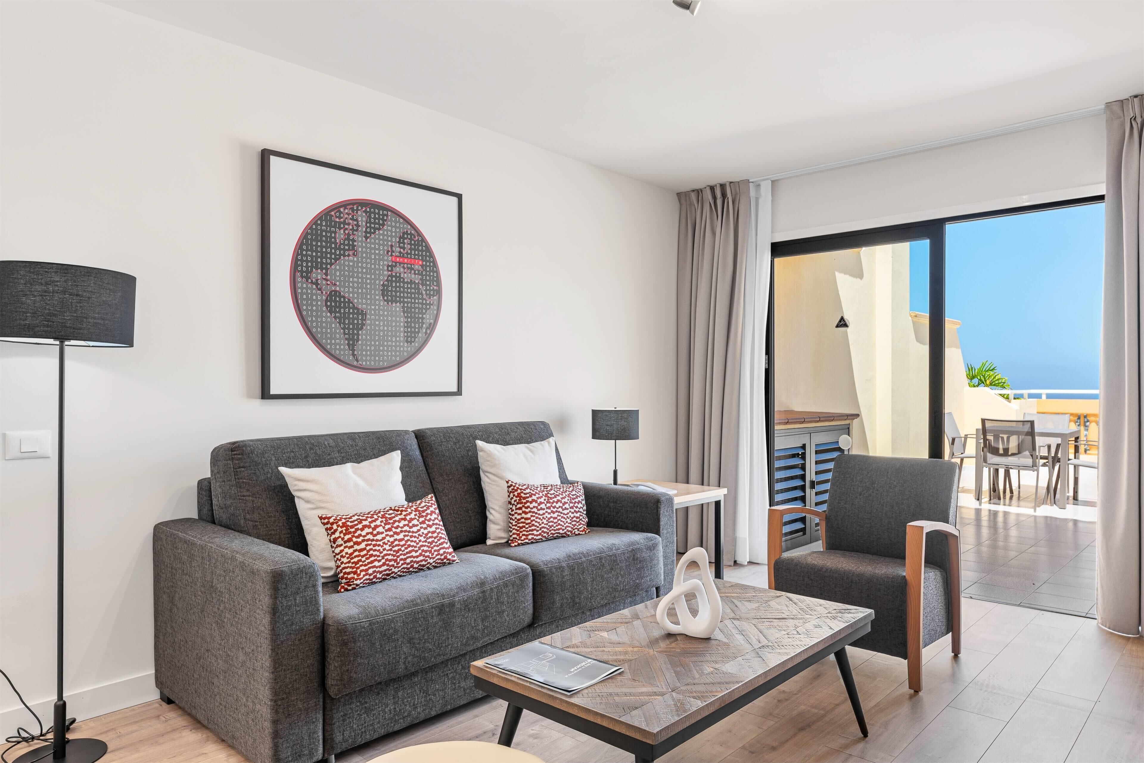 Una sala de estar moderna con un sofá gris, una mesa de centro y un sillón. Hay un ventanal con vistas al mar.