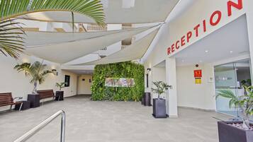 Un área de recepción con un letrero grande, una pared verde con plantas y un espacio de espera con bancos.