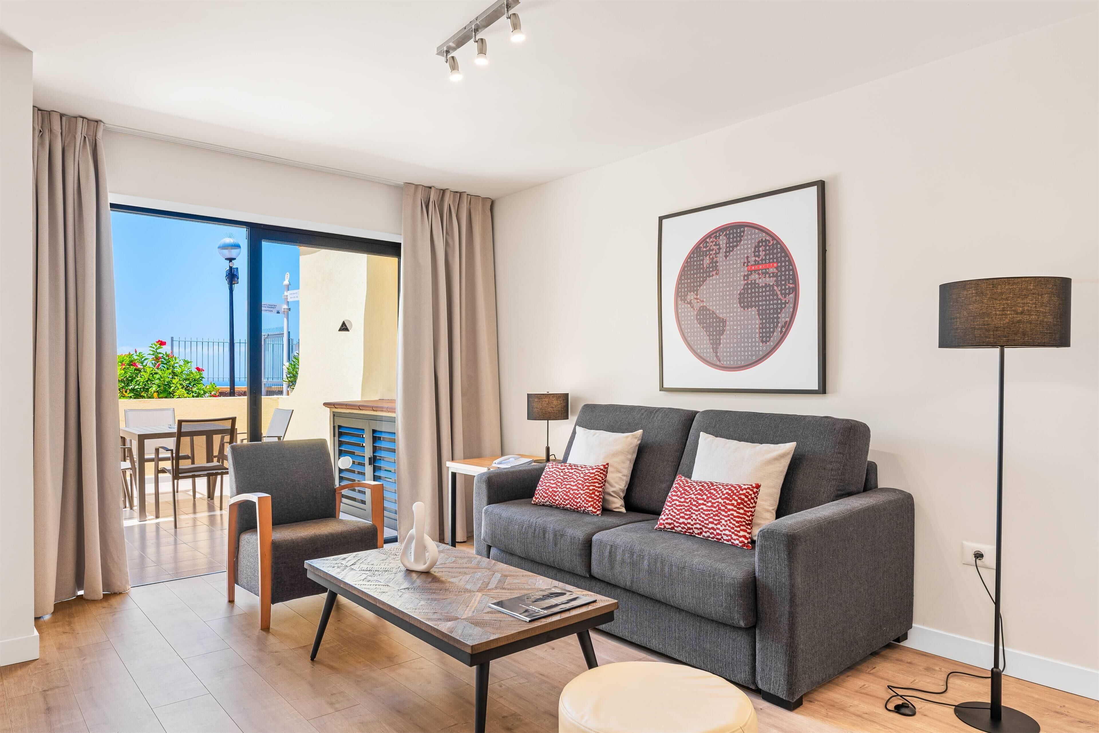 A modern living room with a grey sofa, a coffee table, and a floor lamp. There is a large window with curtains, a view of the outdoors, and a framed artwork on the wall.